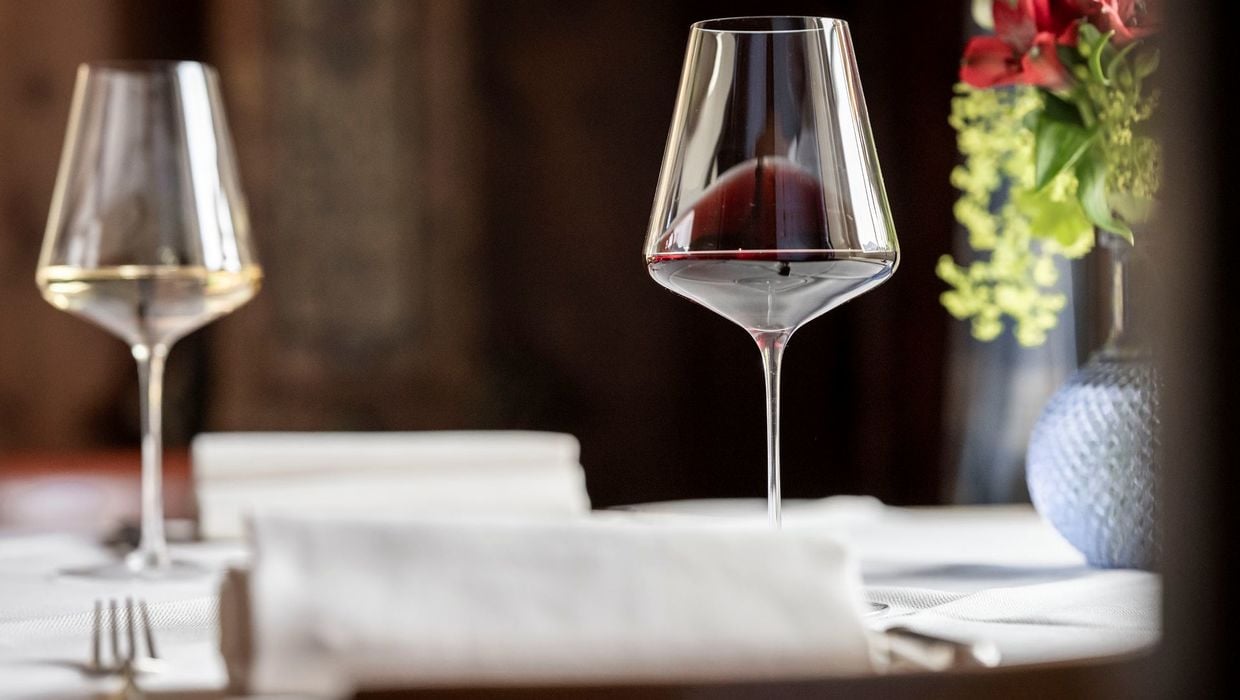 Zwei Weingläser auf einem Tisch, gefüllt mit Weißwein und Rotwein, in einer alten Stube, daneben eine Vase mit Blumen.