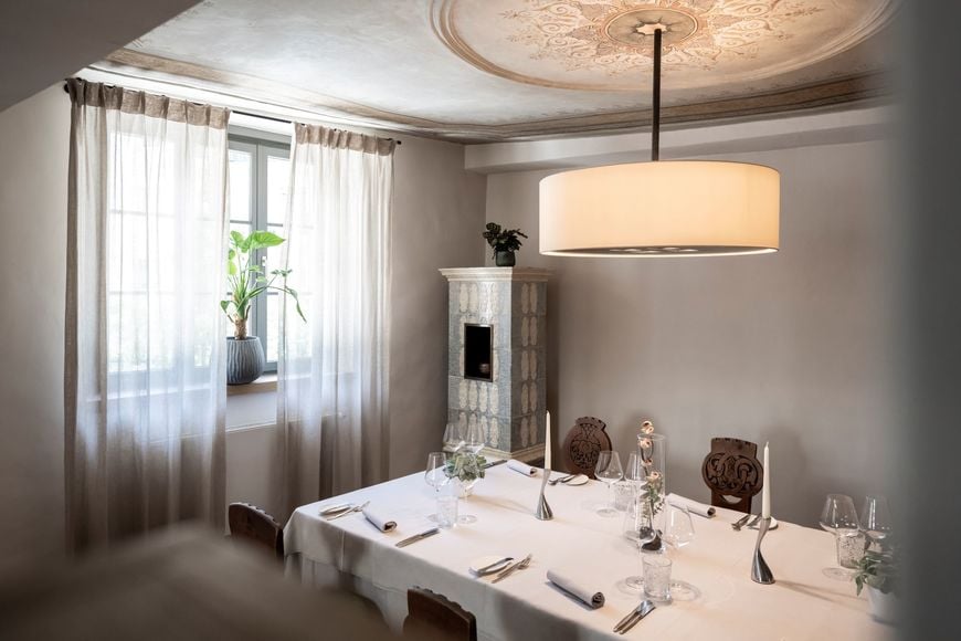 Gedeckter Tisch im Restaurant Löwengrube mit alten Holzstühlen, Pflanze am Fenster, stylischer Lampe und Kachelofen im Hintergrund. ©Löwengrube/Alex Filz