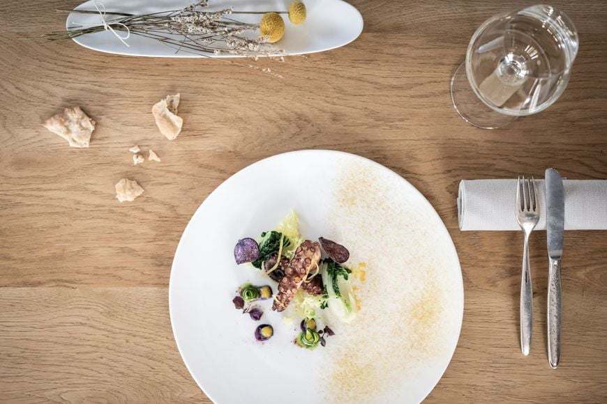 Fischgericht auf Holztisch mit Weißweinglas, Schüttelbrot, Besteck und Blumendekoration. ©Löwengrube/Alex Filz